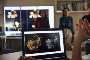 Laptop screen showing a scientific graph and a larger version of the screen behind it, with students surrounding the screens.