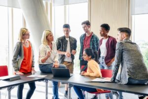 A female teacher speaks to an engaged group of diverse college students gathered around her in class.