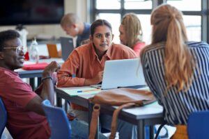 Three students sitting together at a table in a classroom discuss an assignment while one female student uses her laptop.